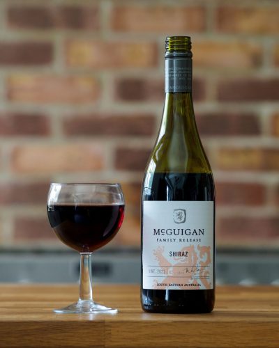 Bottle of McGuigan Family Release Shiraz with filled wine glass on a wooden table against a brick wall.