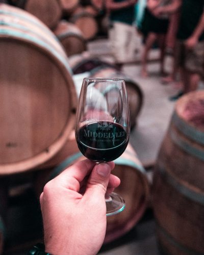 Close-up of a hand holding red wine glass in a Stellenbosch winery with oak barrels in the background.