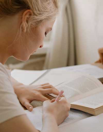 Focused young woman writing notes from an open textbook at a desk, creating a study environment.